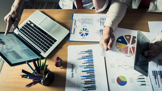 Mehrere Statistiken liegen verteilt auf einem Tisch, mehrere Personen und ein Laptop sind angedeutet