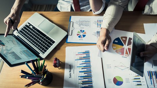 Mehrere Statistiken liegen verteilt auf einem Tisch, mehrere Personen und ein Laptop sind angedeutet