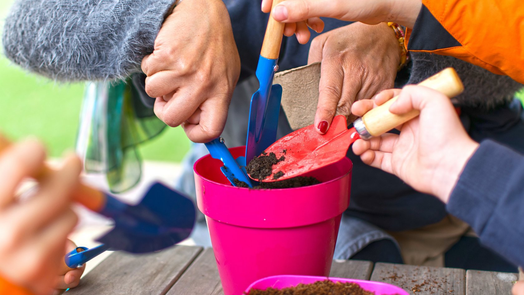 Mehrere Kinderhände geben mit Schaufeln Erde in einen Topf