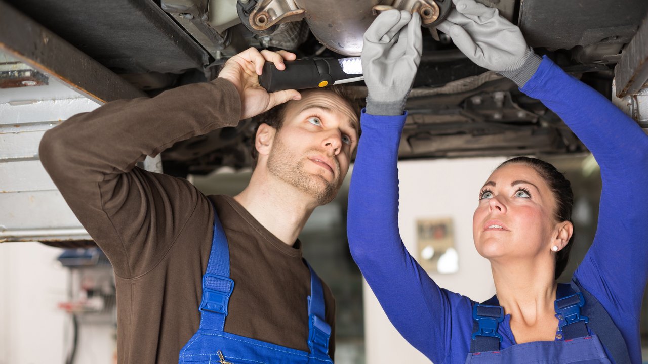 Zwei Mechatroniker (weiblich und männlich) nehmen den Unterboden eines Autos unter die Lupe 