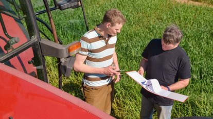 Ein Landwirt und ein Prüfer vor einem Feld und neben einem Traktor im Gespräch