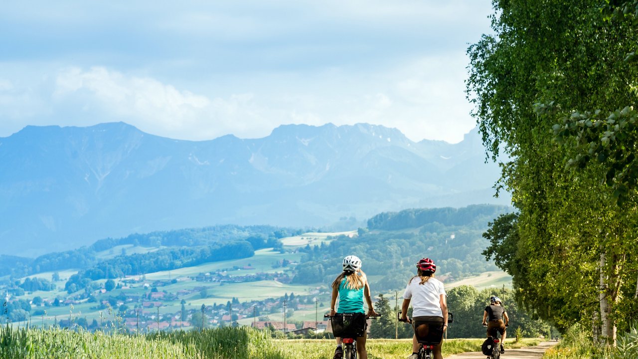 Zwei Radfahrerinnen sind auf einem Fahrradweg unterwegs, im Hintergrund Landschaft mit Bergen