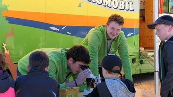 Zwei FÖJler gucken durch Mikroskope, daneben stehen interessierte Kinder