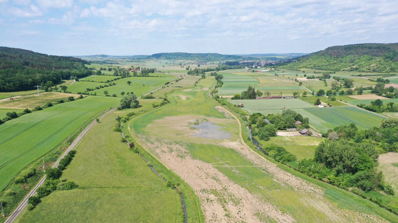 Wiedervernässte Ackerflächen im Ammertal; Bahnlinie links; links und rechts Wald; im Hintergrund blauer bewölkter Himmel