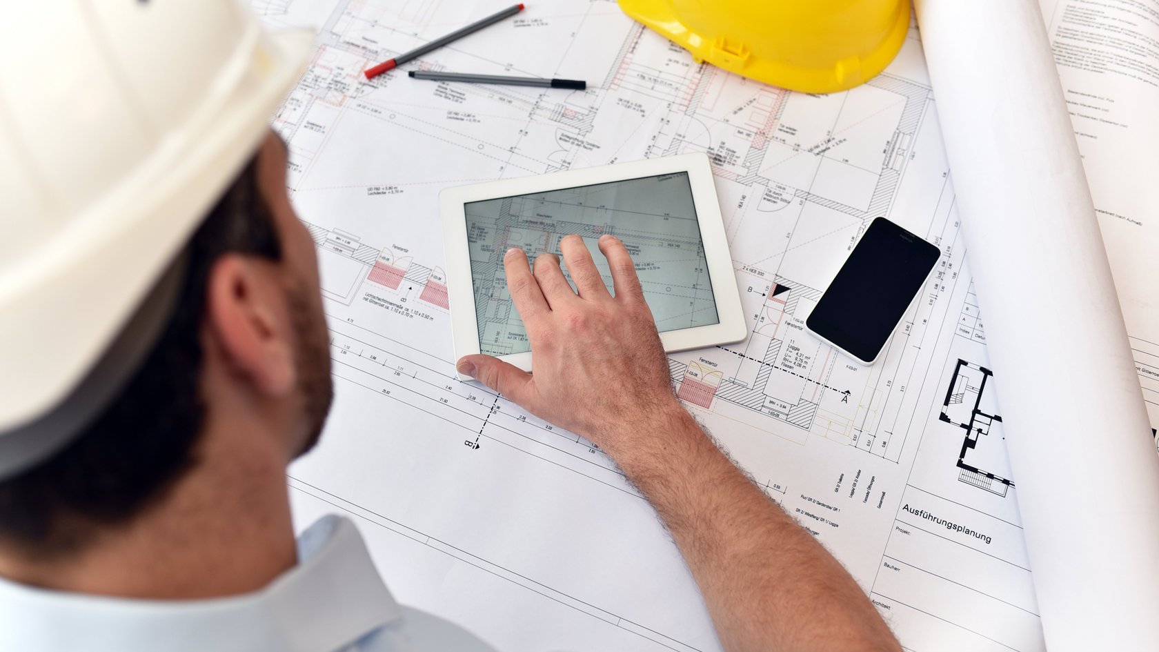 Ein Bauzeichner sitzt mit Tablet, handy und Zeichenutensilien an einem Plan