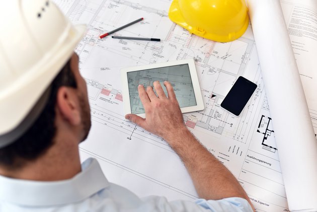 Ein Bauzeichner sitzt mit Tablet, handy und Zeichenutensilien an einem Plan